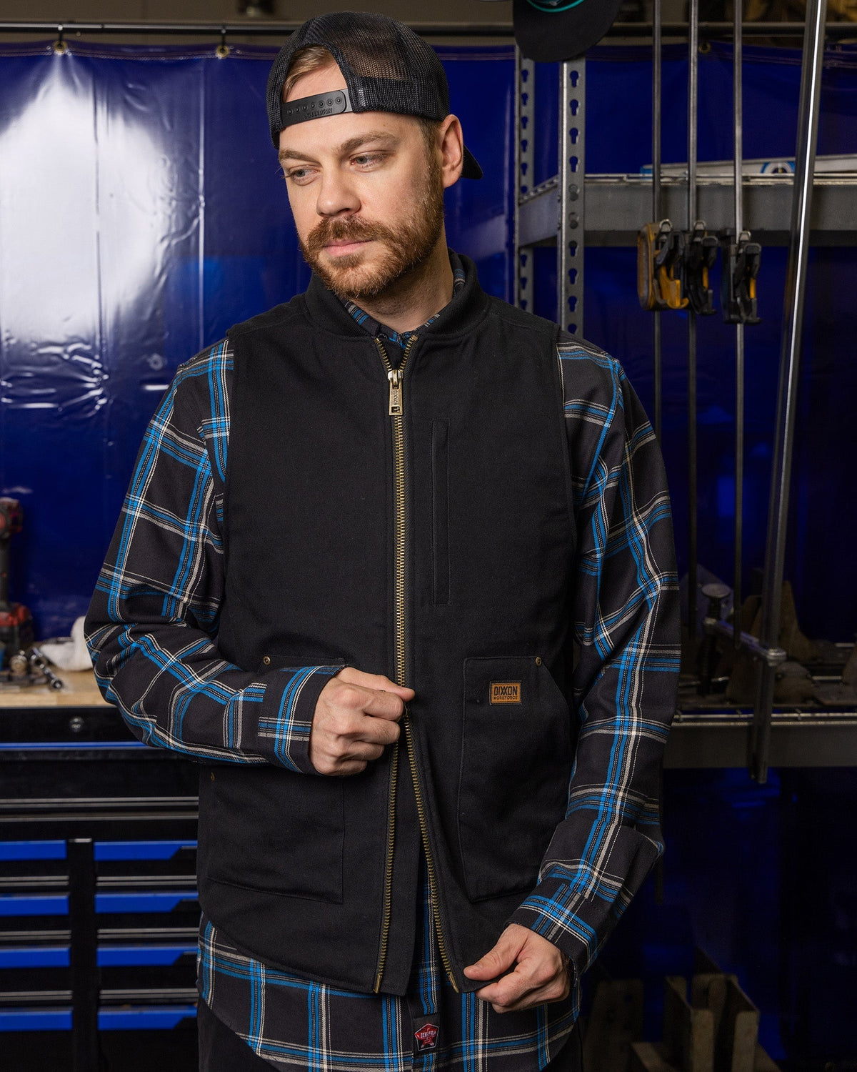 Dixxon Outlaw Work Vest in black worn over blue plaid flannel shirt in workshop setting