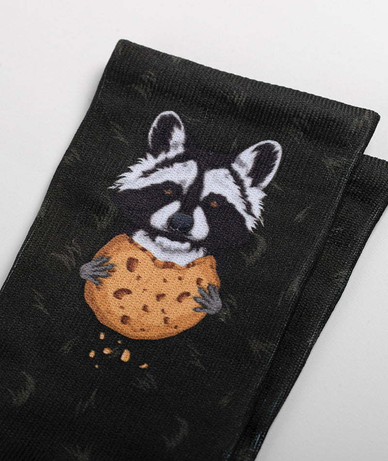 Black crew sock featuring illustrated raccoon holding a large chocolate chip cookie
